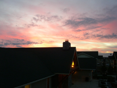 [The light under the eave of the building across the way is the only light except for the expanse of sky which are shades of yellow, peach and white with some scattered small grey wispy clouds.]